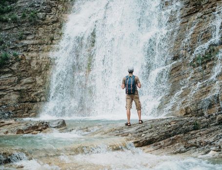 Traveler young man with backpack looking at waterfall in summer outdoor, rear view