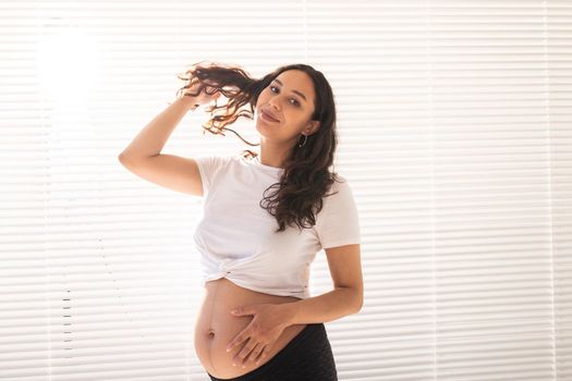 Smiling young beautiful pregnant woman touching her belly and hair and rejoicing. Health and thinking about the future while waiting for baby. Copyspace