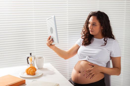 Happy pregnant young beautiful woman talking to mom using video call during morning breakfast. Communication and positive attitude during pregnancy