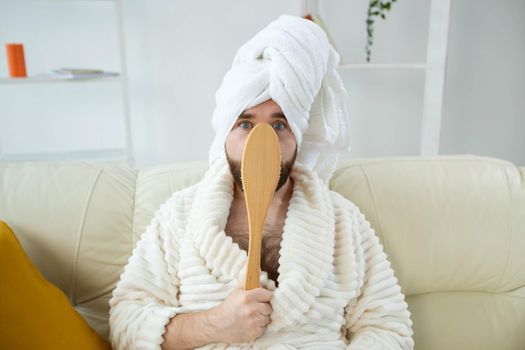 Portrait of cheerful guy wear turban towel holding in hands massage brush while sits on sofa. Male skin care and spa concept