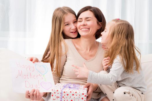 Two little sisters congratulate their mom with Happy mother's day. Children hugging and kissing mother and giving her creative hand-made drawing and gift box