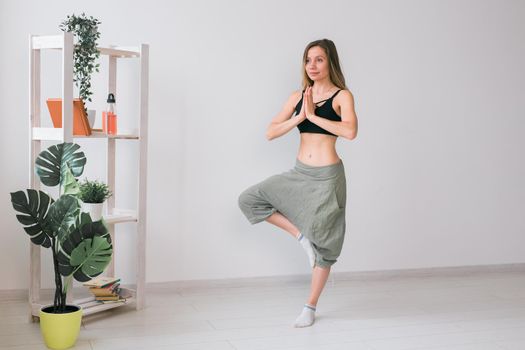 Woman practise yoga performs. Tree pose standing on mat inside of cozy room with plants and greenery. Healthy lifestyle