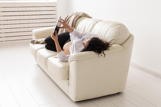 Latin pregnant woman using tablet computer sitting on sofa at home. Pregnancy and information for parenthood