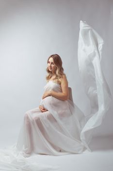 Elegant pregnant young woman standing wearing flying white fabric. Pregnancy, maternity and motherhood.