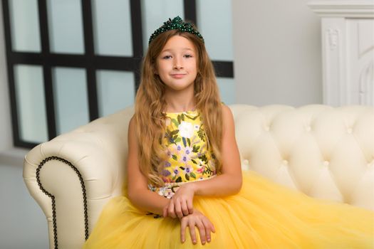 Beautiful little girl in an elegant dress sits on a sofa near a large, panoramic window. Style and fashion concept.