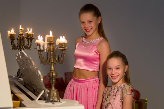 Beautiful Joyful Sisters Posing at White Piano Decorated with Burning Candles in Candlesticks, Portrait of Happy Teenage Girls with Long Hairs Wearing Fashionable Clothes