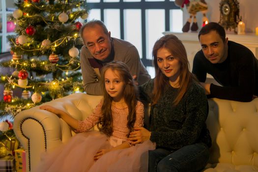 Portrait of loving parents hugging their charming daughter, mom and dad, as well as grandfather sitting on the couch near the Christmas Tree. Against the background of a large panoramic window in the studio. Holidays concept.