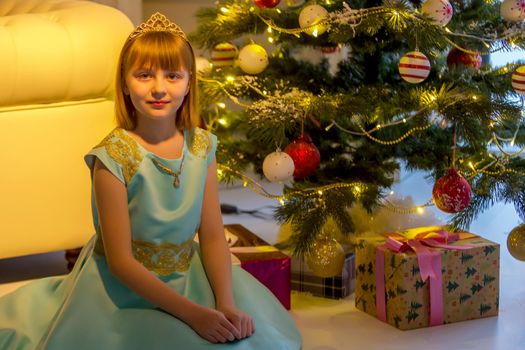 Happy little girl rejoices at Christmas present. A girl sits near the shining lights of a Christmas tree which stands near a large panoramic window.