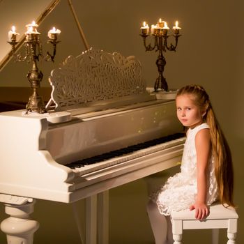 Beautiful little girl is playing on a white piano in a dark room by candlelight. The concept of Christmas and New Year, family happiness.