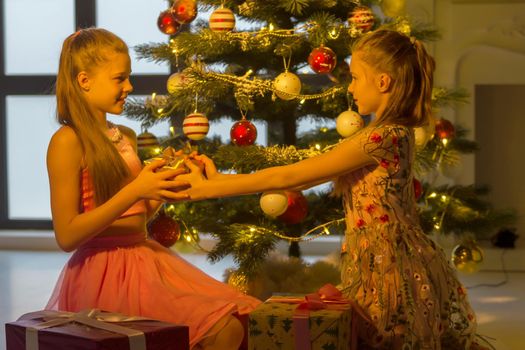 Two Beautiful Smiling Teen Girls Sitting on the Floor Looking at Each Other in front of Christmas Tree, Two Adorable Sisters Wearing Nice Dresses Exchanging Gifts at Christmas Warm Cozy Room Interior.