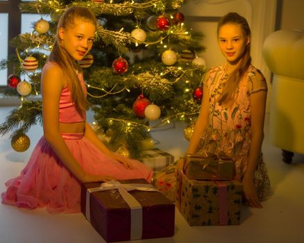 Beautiful Smiling Sisters Sitting on their Knees Opposite Each Other at Christmas Tree, Two Adorable Teen Girls Wearing Nice Dresses Exchanging Gifts at Christmas Warm Cozy Room Interior.