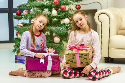 Happy Girls in Pajamas Sitting on the Floor with Gift Boxes in Front of Decorated Christmas Tree, Two Cheerful Sisters Looking at Camera, Happy Family Celebrating Xmas and New Year
