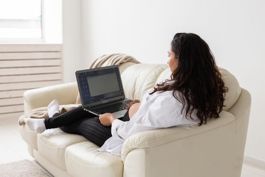 Latin pregnant woman using laptop sitting on sofa at home. Pregnancy and information for parenthood