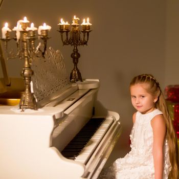 Beautiful little girl is playing on a white piano in a dark room by candlelight. The concept of Christmas and New Year, family happiness.