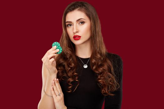 Pretty long hair woman in black dress holding chips for gambling in casino. Studio shot on red background. Poker. Two green Chips in Hand