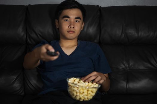 young man holding remote control and watching TV while sitting on sofa at night