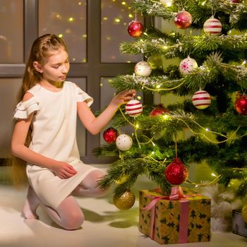 Adorable little girl decorating the Christmas tree with colorful glass baubles. Trimming the Christmas tree. Celebrating Xmas at home.