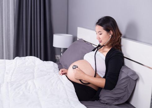 Pregnant woman listening to her belly with a stethoscope on the bed