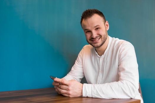 A young attractive European businessman in a white shirt and headphones spends time in a smartphone, sitting at a table in a cafe on a coffee break.