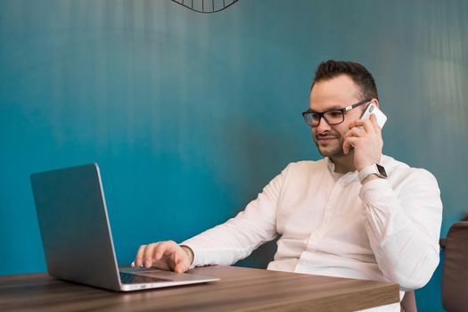 Young businessman in glasses and a white shirt talking on the phone and sitting in a laptop on a blue background.