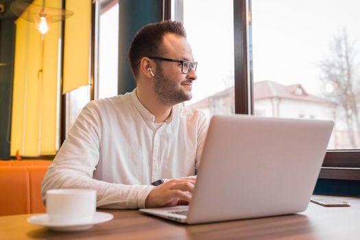 Young satisfied businessman sits in a laptop and looks out the window on a background of cafe interior.