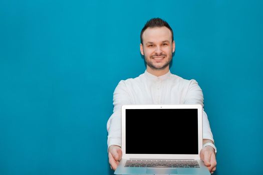 Young attractive businessman with a beard smiles and holds a laptop mock up on a blue background.