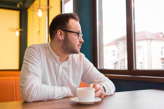 Young attractive businessman in a white shirt, glasses and headphones drinks coffee and looks thoughtfully out the window.