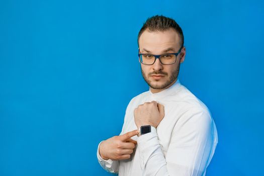 Portrait of young attractive european businessman in white shirt, glasses and beard pointing with hand at his smartwatch on blue background.