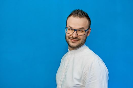 Portrait of a young attractive european businessman in a white shirt, glasses, with a beard on a blue background.