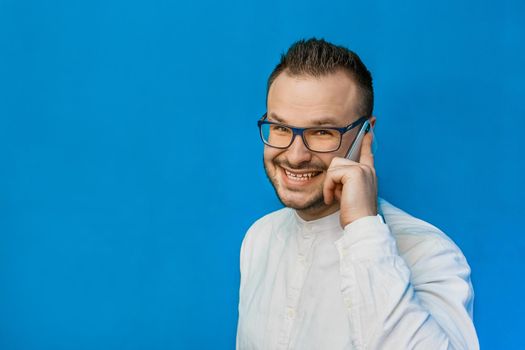 Young attractive businessman in a white shirt and glasses speaks on the phone and smiles on a blue background.