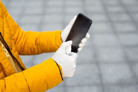 Young woman disinfects her phone with a hand sanitizer. Woman holding a mobile phone in her hands wearing latex protective gloves at the street.