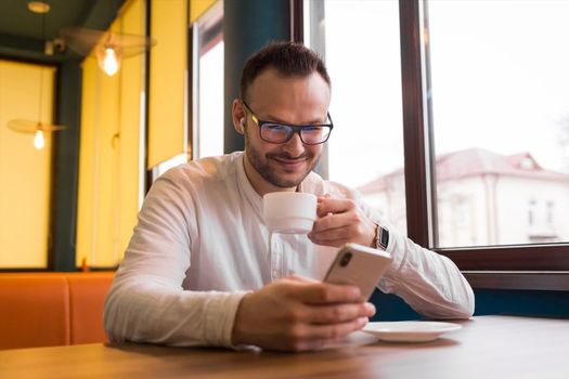 Portrait of a young handsome businessman in a white shirt, glasses, with a beard and headphones, spending time on the phone, drinking coffee and smiling.
