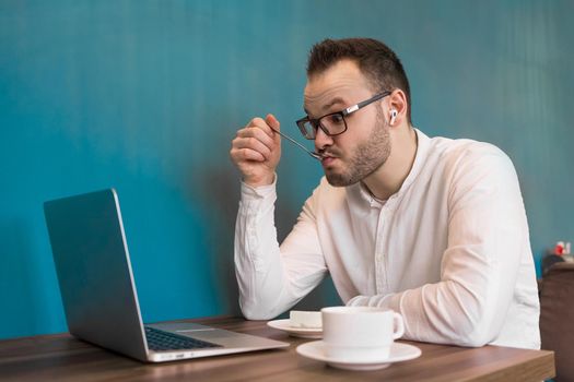 Attractive businessman in a white shirt looks in surprise at a laptop and drinks coffee on a blue background.