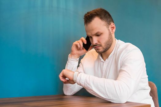 A young European guy businessman in a white shirt, with a beard sits at a table, talks on a mobile phone and looks at the time on a blue background.