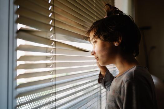 Nine-years-old girl looking out the window through the blinds in the hope of being released from quarantine due to the Covid-19 epidemic
