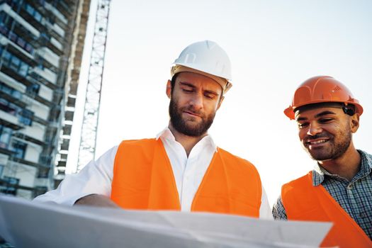 Two young male engineers in uniform and hardhats working at construction site, close up