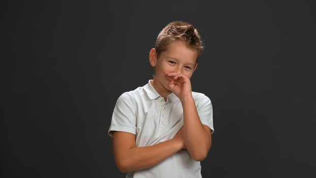 Shyly laughing boy covers his face with his hand looking at the camera wearing white polo shirt and black pants isolated on black background.