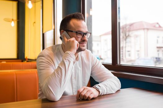 Young attractive businessman in a white shirt and glasses sits at a table in a cafe and talking on the phone, smiling.