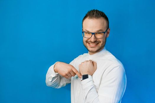 Portrait of young attractive european businessman in white shirt, glasses and beard pointing with hand at his smartwatch on blue background.