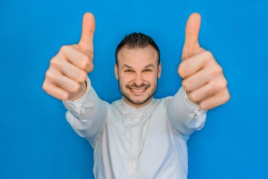 Portrait of young attractive european happy businessman in white shirt with beard showing cool thumbs up two hands on blue background.