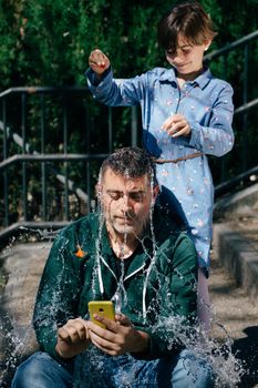 Little girl blowing up a water-filled balloon over her father's head to get him to stop staring at his smartphone.