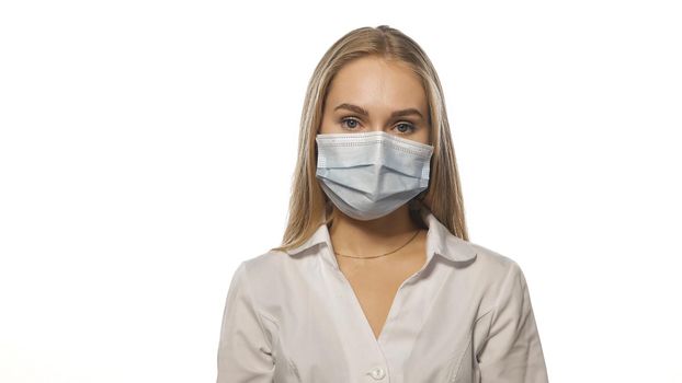 Nurse in a medical mask and white uniform with blond loose hair looking at the camera. Isolated on white background.