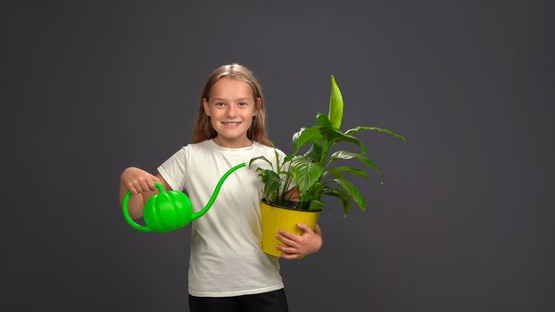 Little girl watering a flower in pot using a funny watering can while holding everything in her hands. Girl wearing white t shirt smiling at camera. Isolated on dark grey or black background.