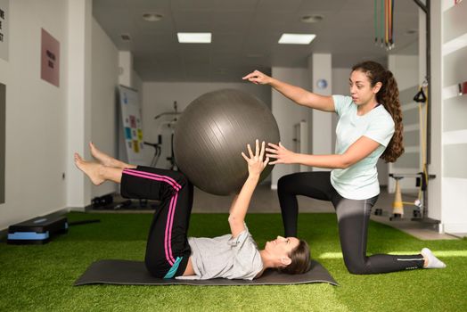 Physical therapist assisting young caucasian woman with exercise with yoga ball during rehabilitation in the gym at hospital. Female physiotherapist training a patient in physiotherapy center.