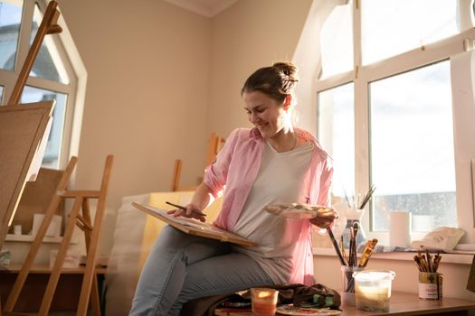 Caucasian woman artist working on a painting in bright daylight studio. Happy artist draws an art project with paints and a brush in the workshop. Hobby. Artist at work. Creative profession.