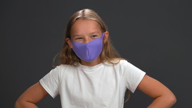 School girl wearing blue reusable face mask wearing white t-shirt smiling with her eyes looking at camera while standing in studio. Isolated on black background. Kids fashion concept.