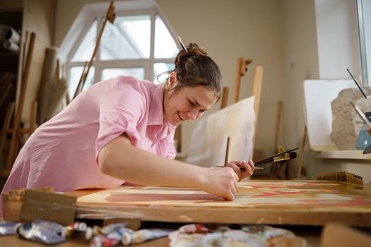 Caucasian woman artist working on a painting in bright daylight studio. Happy artist draws an art project with paints and a brush in the workshop. Hobby. Artist at work. Creative profession.