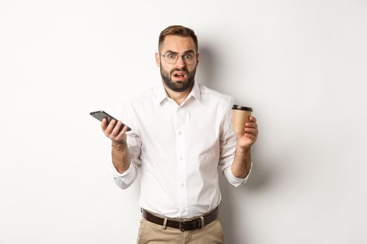 Image of man drinking coffee, feeling confused about strange message on mobile phone, standing over white background.