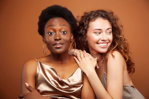 two pretty girls african and caucasian blond posing cheerful together on brown background, ethnicity diverse lifestyle people concept closeup