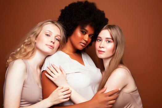 young pretty caucasian, afro, scandinavian woman posing cheerful together on brown background, lifestyle diverse nationality people concept closeup